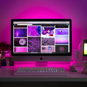 A desktop setup with a pink-lit computer display showing pink and purple-themed images, surrounded by a candle, camera, photo frame, and mug.