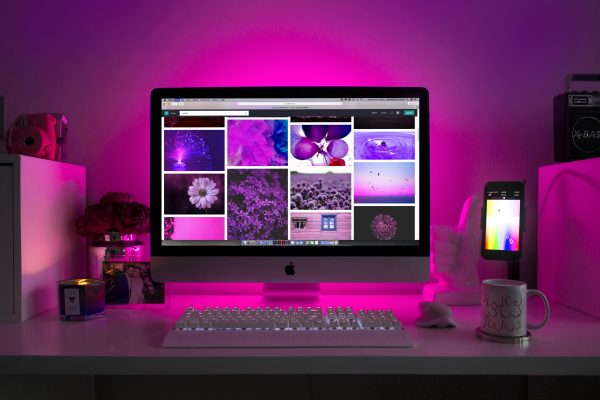 A desktop setup with a pink-lit computer display showing pink and purple-themed images, surrounded by a candle, camera, photo frame, and mug.